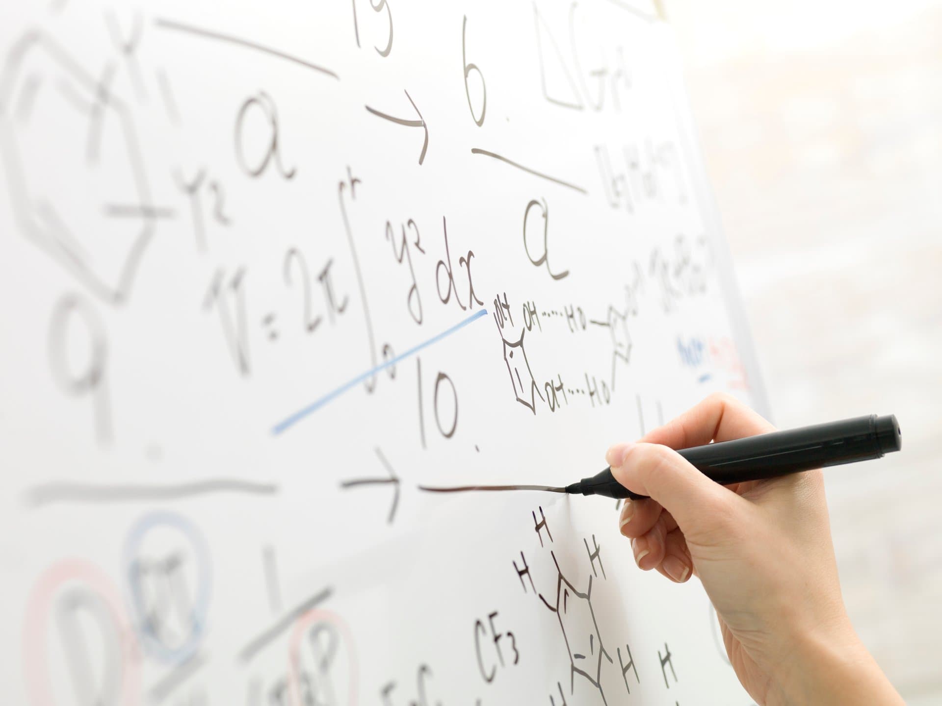 Close-up of a hand writing mathematical equations and chemical structures on a whiteboard with a black marker during clinical research.