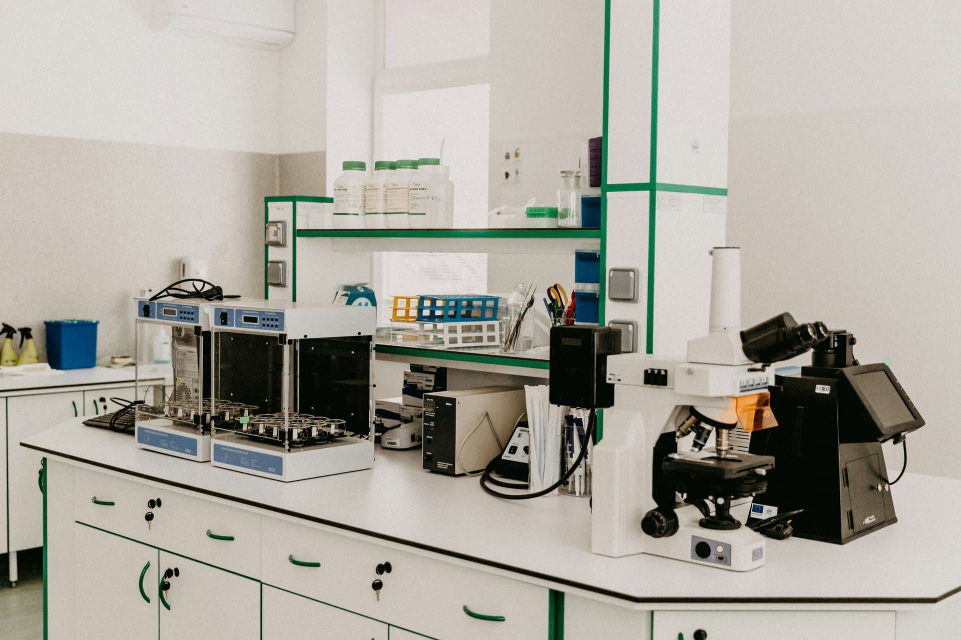 Bright clinical laboratory featuring a microscope, lab incubators, test tube racks, and reagent bottles on white workbenches with green trim.