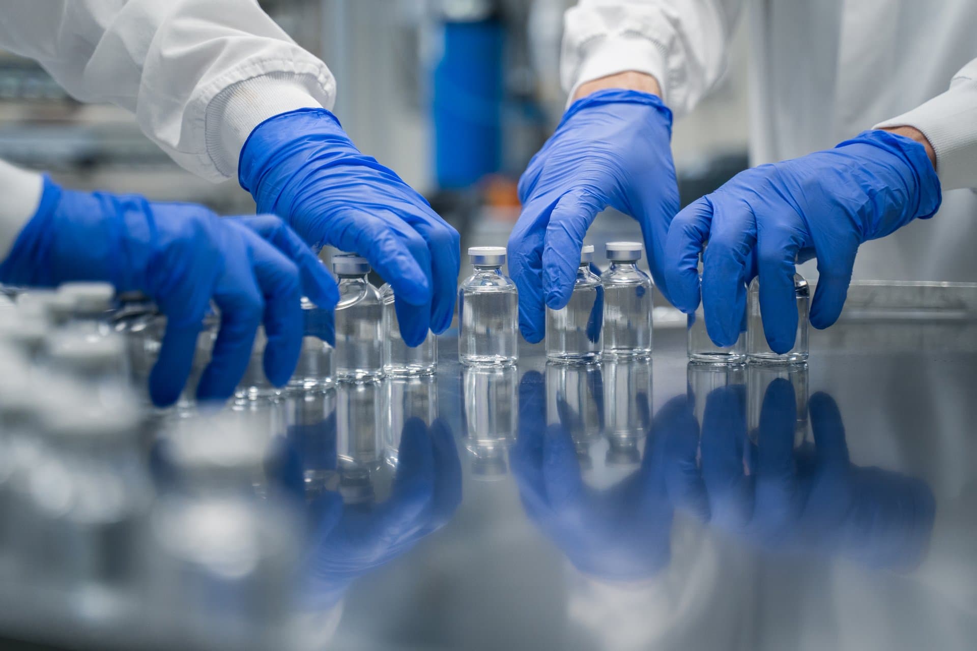 Close-up of two healthcare workers wearing blue nitrile gloves arranging clear glass medication vials on a reflective lab surface.