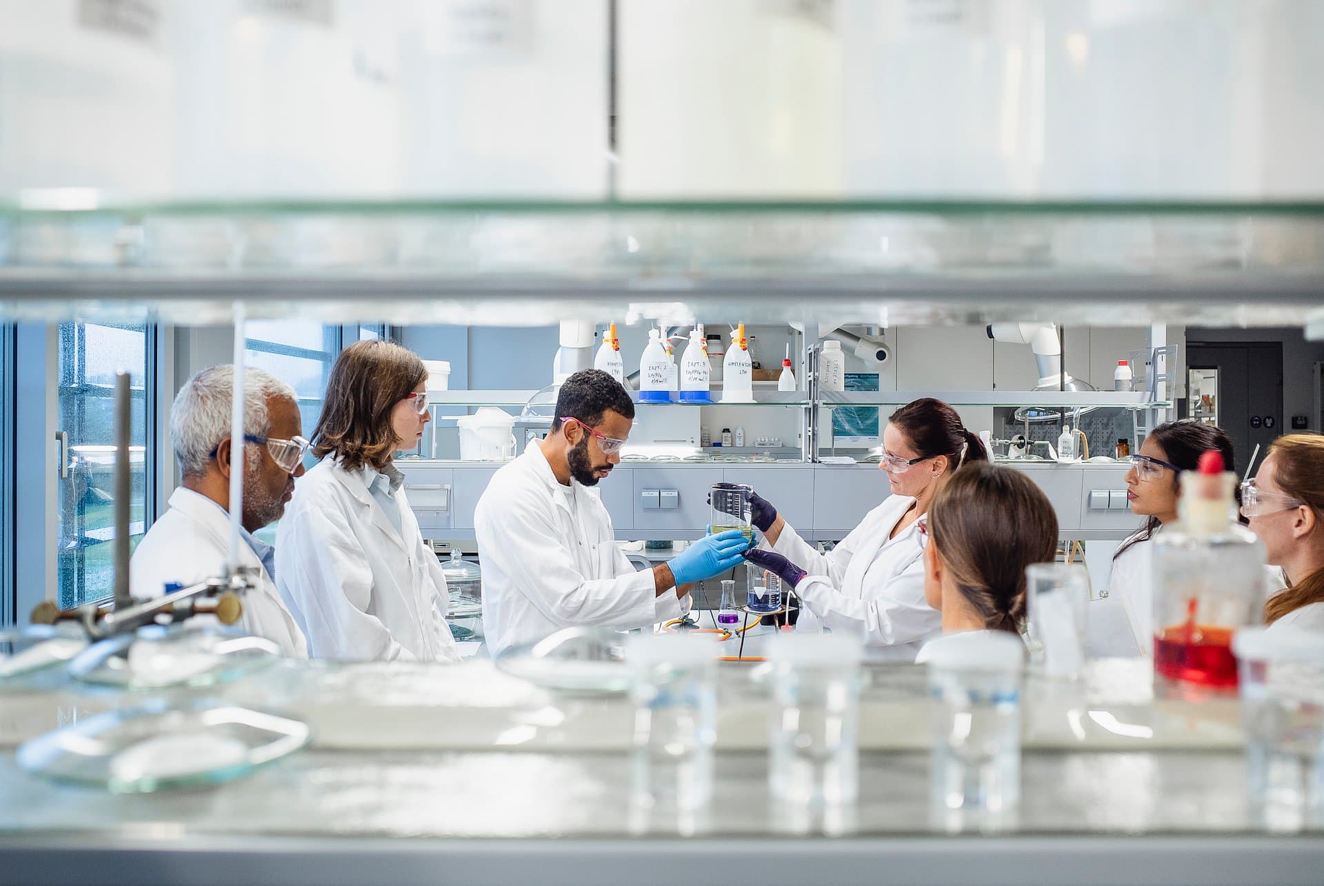 Group of scientists in white coats and safety goggles examining beaker of liquid together in a bright clinical research laboratory setting.